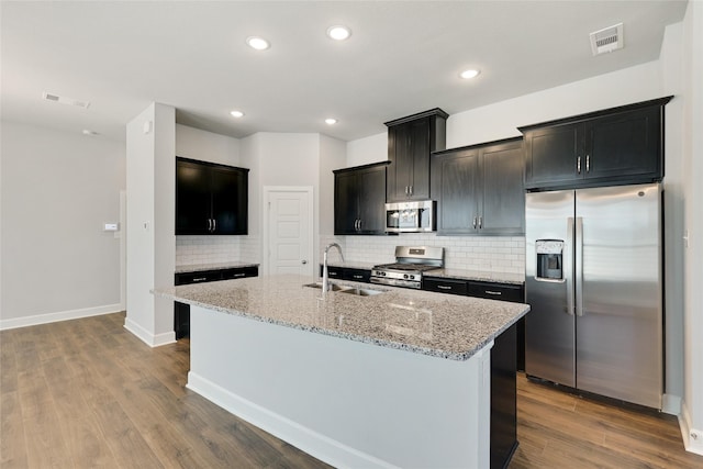 kitchen with hardwood / wood-style floors, sink, a kitchen island with sink, stainless steel appliances, and light stone countertops