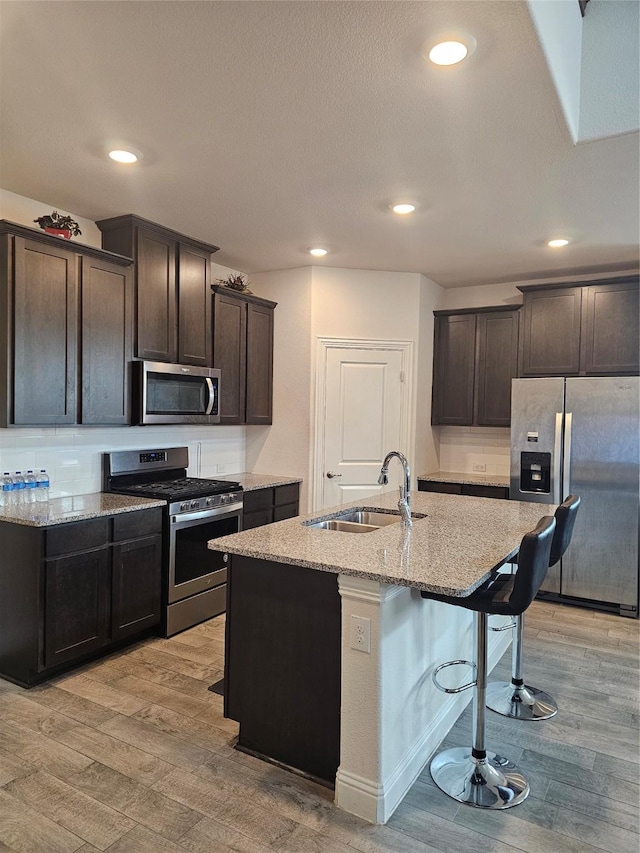 kitchen featuring sink, appliances with stainless steel finishes, light stone countertops, a center island with sink, and light wood-type flooring