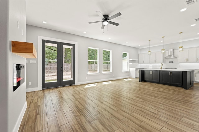 kitchen featuring a kitchen island with sink, hanging light fixtures, white cabinets, french doors, and wall chimney exhaust hood