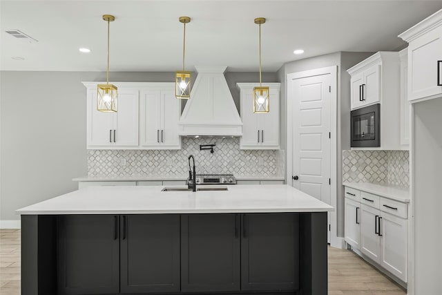 kitchen featuring an island with sink, sink, white cabinets, custom exhaust hood, and hanging light fixtures