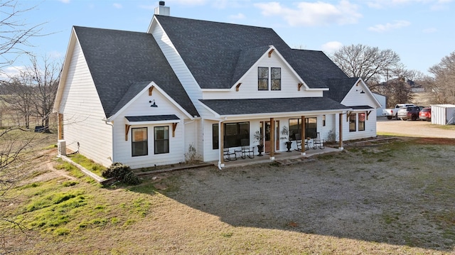 modern farmhouse with a shingled roof, a chimney, a front lawn, and a porch