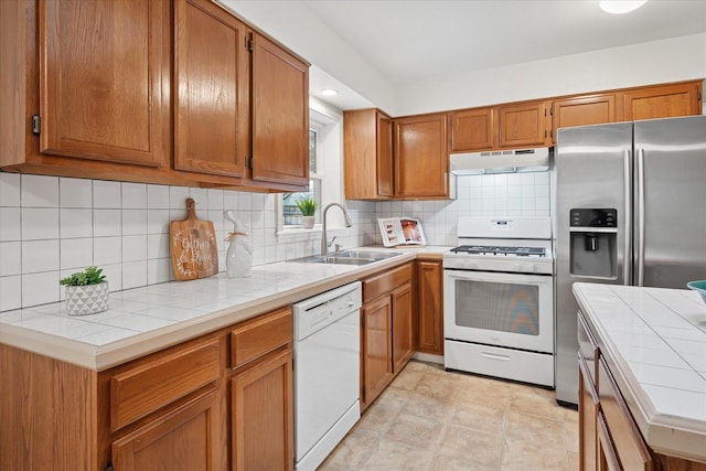 kitchen with tile countertops, under cabinet range hood, white appliances, a sink, and brown cabinets