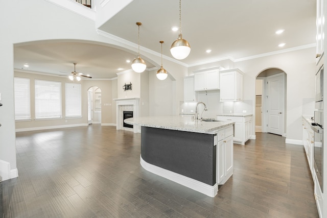 kitchen with ceiling fan, hanging light fixtures, light stone countertops, an island with sink, and white cabinets