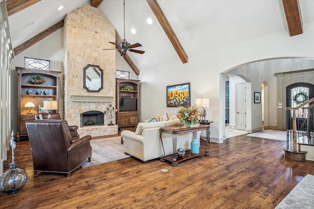 living area featuring arched walkways, stairway, wood finished floors, a stone fireplace, and beam ceiling