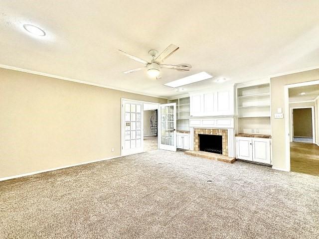 unfurnished living room featuring crown molding, ceiling fan, carpet flooring, a brick fireplace, and built in shelves