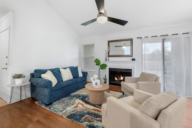 living area with a warm lit fireplace, ceiling fan, wood finished floors, vaulted ceiling, and crown molding