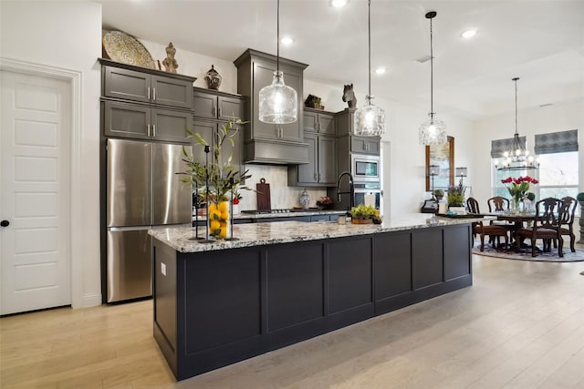 kitchen with appliances with stainless steel finishes, hanging light fixtures, a large island, light stone counters, and light hardwood / wood-style floors