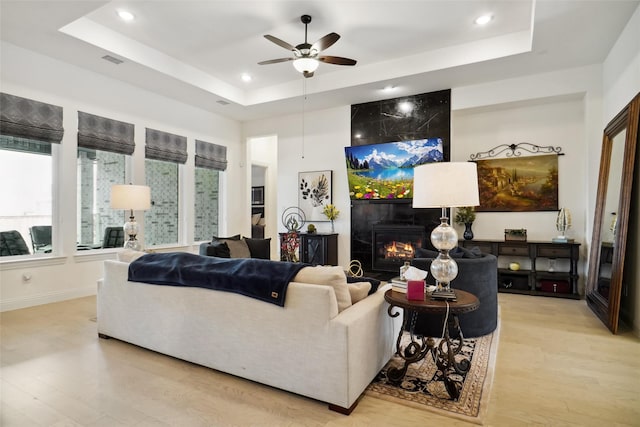 living room featuring ceiling fan, a fireplace, a raised ceiling, and light wood-type flooring