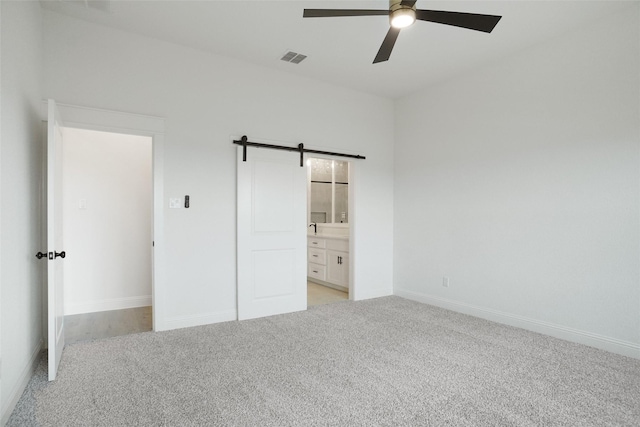 unfurnished bedroom featuring ceiling fan, a barn door, ensuite bath, and light carpet