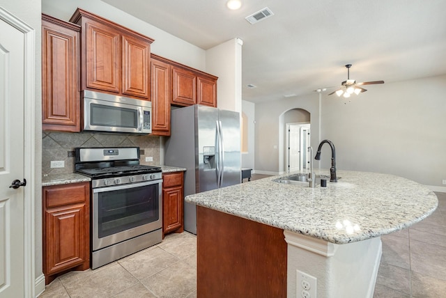 kitchen with appliances with stainless steel finishes, sink, backsplash, a kitchen island with sink, and light stone countertops