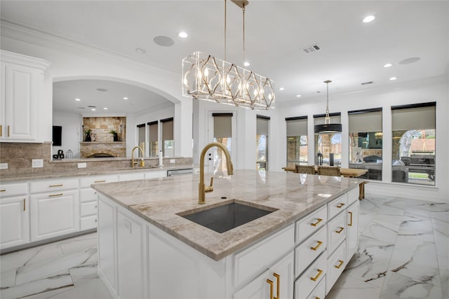 kitchen featuring an island with sink, white cabinetry, sink, hanging light fixtures, and light stone counters