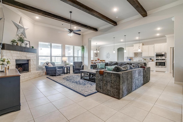 tiled living room featuring beam ceiling, a stone fireplace, ceiling fan with notable chandelier, and ornamental molding