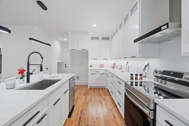 kitchen featuring wall chimney range hood, sink, hanging light fixtures, stainless steel appliances, and white cabinets