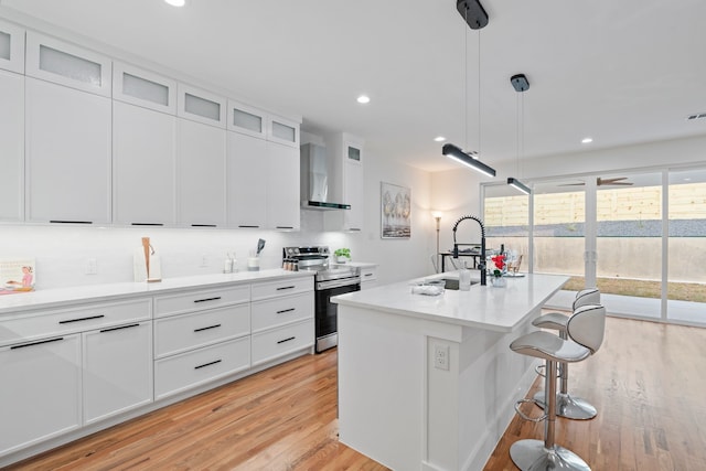 kitchen featuring a kitchen island with sink, wall chimney range hood, stainless steel electric range, and white cabinetry