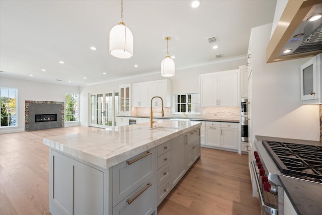 kitchen with range hood, light stone counters, high end stove, decorative light fixtures, and a large island with sink