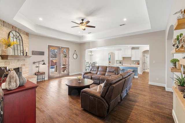 living room featuring french doors, a stone fireplace, a raised ceiling, and ceiling fan with notable chandelier