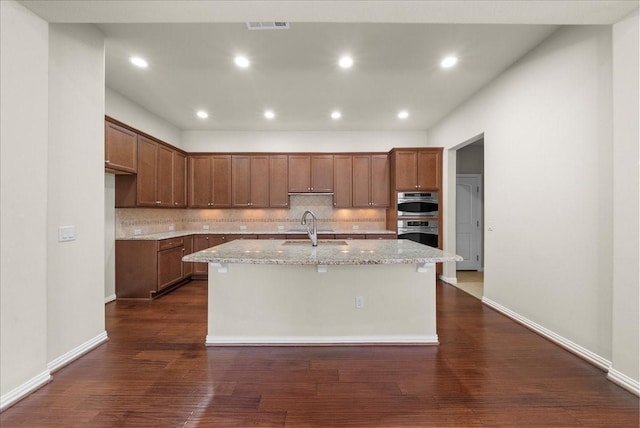 kitchen with tasteful backsplash, visible vents, dark wood-style flooring, a kitchen island with sink, and a sink