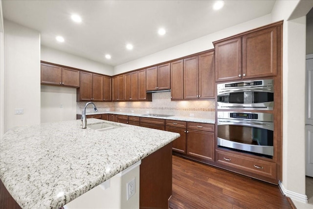 kitchen with decorative backsplash, dark wood-style floors, light stone countertops, double oven, and a sink