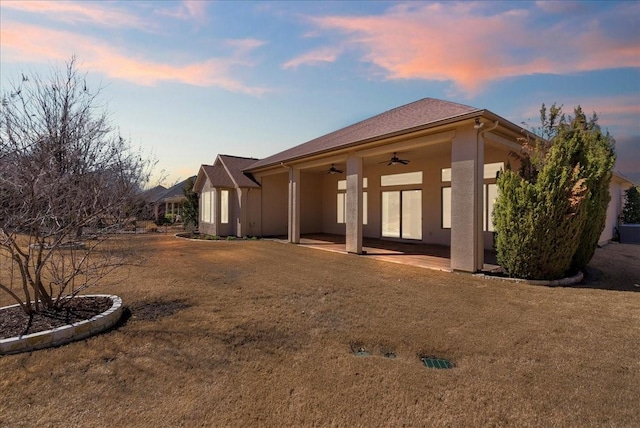 back of house at dusk with a patio area, ceiling fan, a lawn, and stucco siding