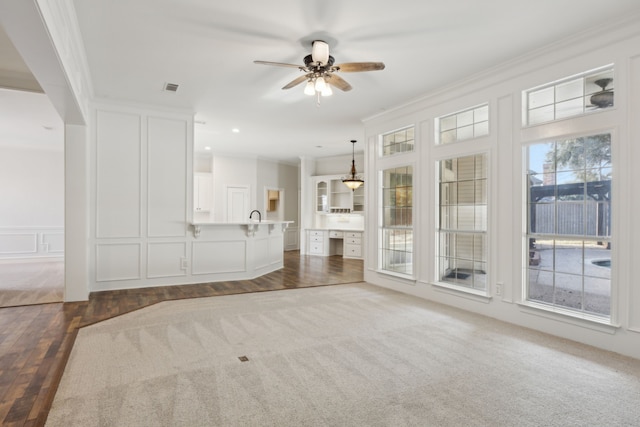 unfurnished living room with a wealth of natural light, ornamental molding, ceiling fan, and dark colored carpet