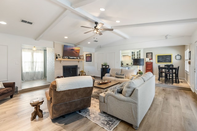 living room with ceiling fan, a fireplace, light wood-type flooring, and vaulted ceiling with beams