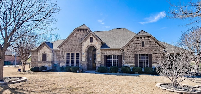 french country home with stone siding, roof with shingles, a front lawn, and brick siding