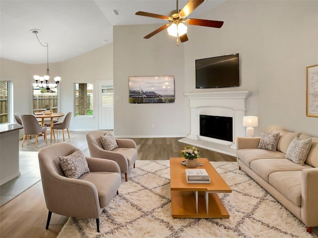 living room with baseboards, high vaulted ceiling, ceiling fan with notable chandelier, light wood-style floors, and a fireplace with raised hearth