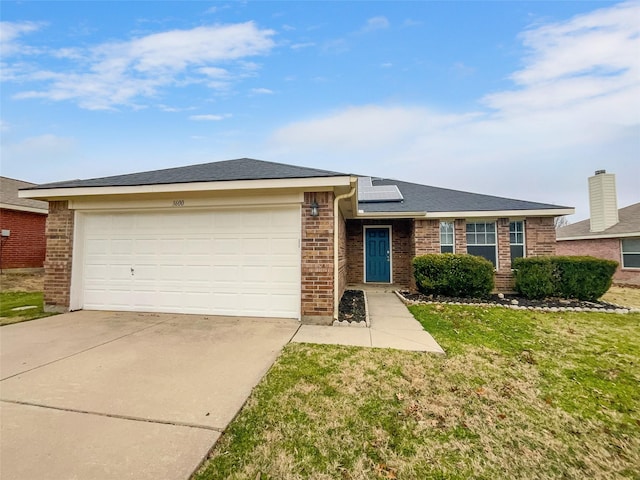 ranch-style home featuring driveway, brick siding, an attached garage, roof mounted solar panels, and a front yard