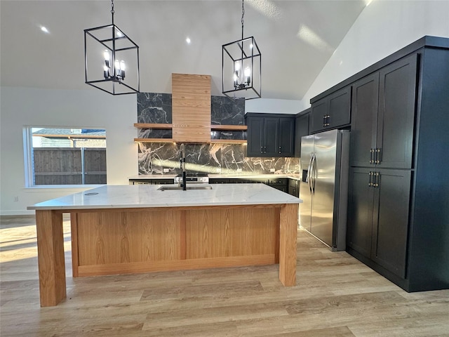 kitchen with light stone counters, light wood-type flooring, pendant lighting, stainless steel fridge, and a kitchen island with sink