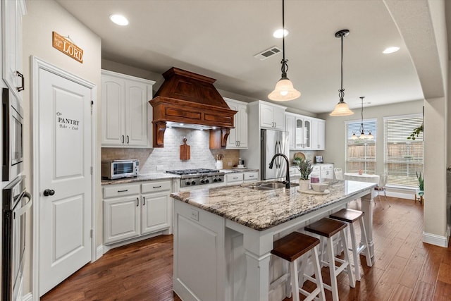 kitchen with custom range hood, decorative light fixtures, stainless steel appliances, white cabinets, and a kitchen island with sink