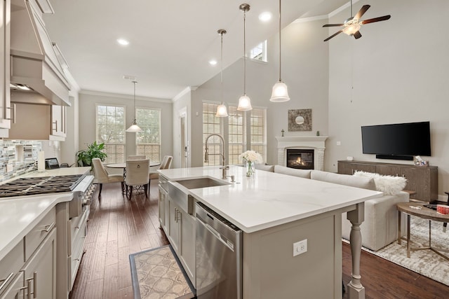 kitchen with a center island with sink, custom exhaust hood, hanging light fixtures, appliances with stainless steel finishes, and a sink