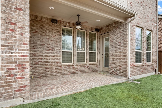 view of exterior entry featuring ceiling fan, a patio, brick siding, and a lawn