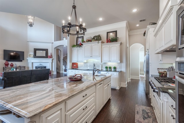 kitchen featuring arched walkways, decorative light fixtures, white cabinetry, a kitchen island with sink, and light stone countertops