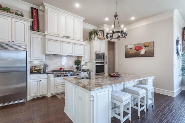 kitchen with a center island with sink, light stone counters, built in appliances, hanging light fixtures, and white cabinetry