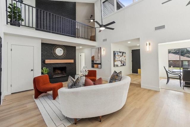 living room with a brick fireplace, light wood-type flooring, ceiling fan, and a towering ceiling