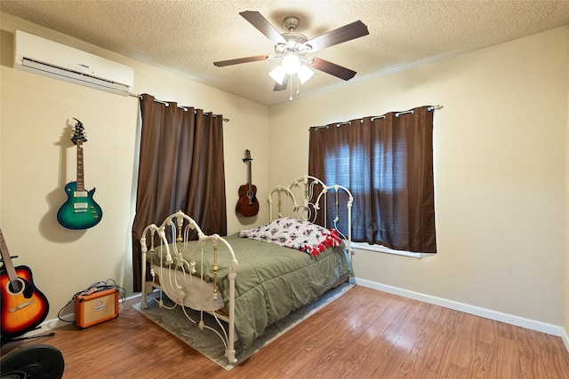 bedroom featuring a ceiling fan, wood finished floors, baseboards, a wall unit AC, and a textured ceiling