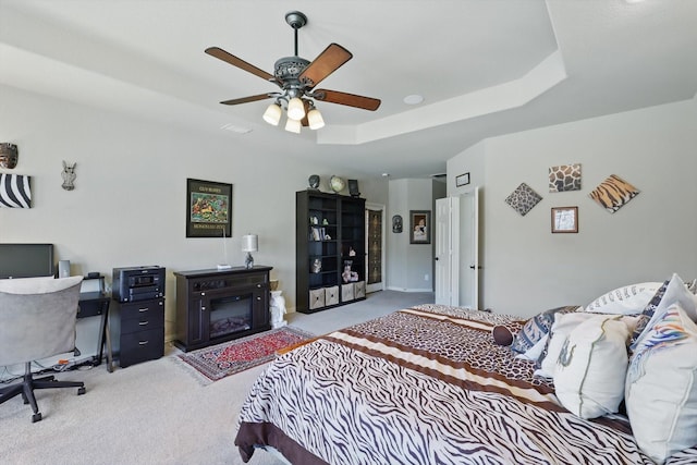 carpeted bedroom featuring a glass covered fireplace, a tray ceiling, and ceiling fan