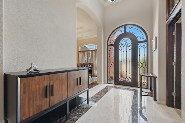 foyer entrance featuring arched walkways, a textured wall, baseboards, marble finish floor, and ornamental molding