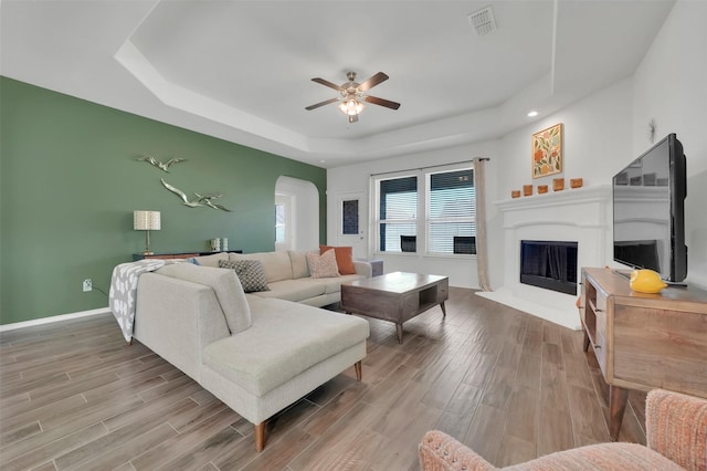 living room featuring arched walkways, a fireplace with raised hearth, wood finished floors, and a raised ceiling