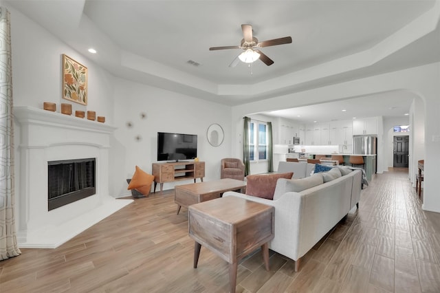 living area with arched walkways, a fireplace with raised hearth, visible vents, light wood-type flooring, and a tray ceiling