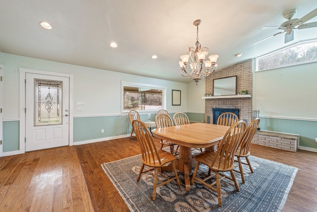 dining room featuring hardwood / wood-style flooring, vaulted ceiling, a brick fireplace, and ceiling fan with notable chandelier