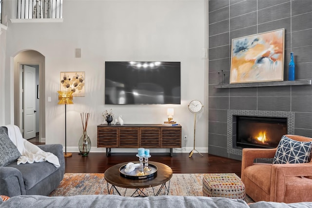 living room featuring arched walkways, dark wood-style flooring, a tiled fireplace, and baseboards