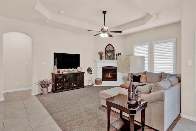 living room featuring light tile patterned floors, arched walkways, a raised ceiling, a ceiling fan, and a glass covered fireplace