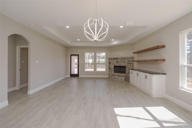 unfurnished living room with light wood-type flooring, a chandelier, a stone fireplace, and a raised ceiling