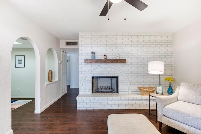 living room featuring a ceiling fan, visible vents, baseboards, a brick fireplace, and dark wood-style floors