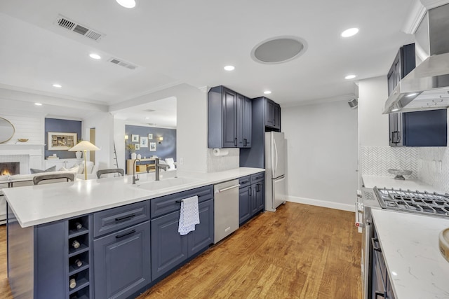 kitchen featuring blue cabinets, appliances with stainless steel finishes, wall chimney range hood, crown molding, and a breakfast bar area