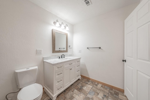 bathroom with toilet, vanity, visible vents, baseboards, and stone finish flooring