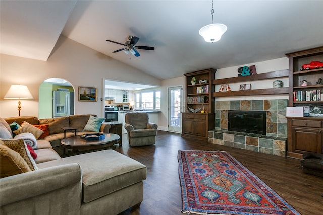 living room with ceiling fan, arched walkways, a tile fireplace, dark wood-style flooring, and vaulted ceiling