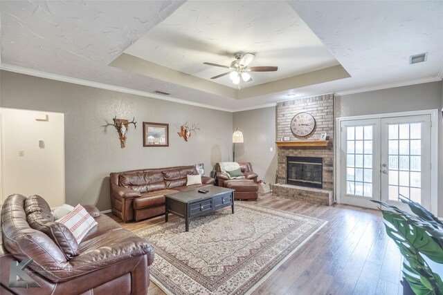 living room featuring a brick fireplace, light hardwood / wood-style floors, crown molding, ceiling fan, and a raised ceiling