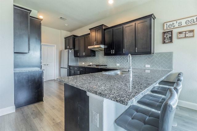 kitchen featuring light stone counters, freestanding refrigerator, under cabinet range hood, stainless steel gas stovetop, and a sink